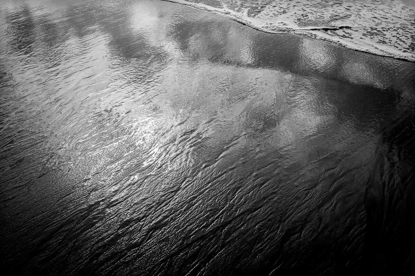 Black and white photograph of reflective tidal water on the Maine coast, where retreating waves mirror clouds and sky across wet sand.