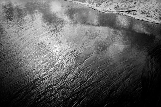 Black and white photograph of reflective tidal water on the Maine coast, where retreating waves mirror clouds and sky across wet sand.
