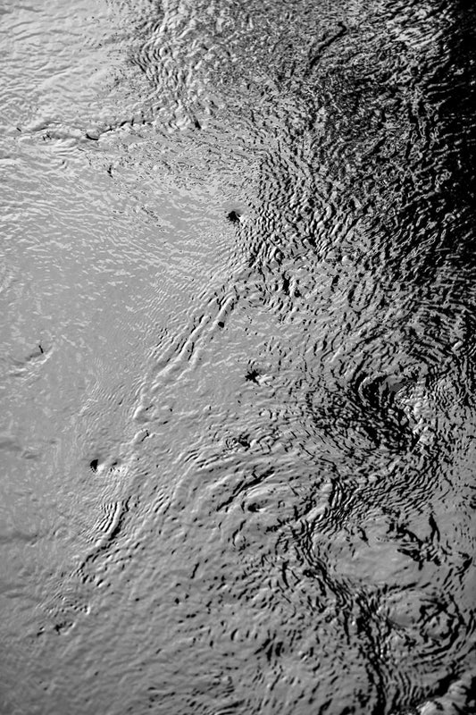 Black and white photograph of swirling water currents in a Maine stream, showing abstract patterns, eddies, and reflective surface movement.