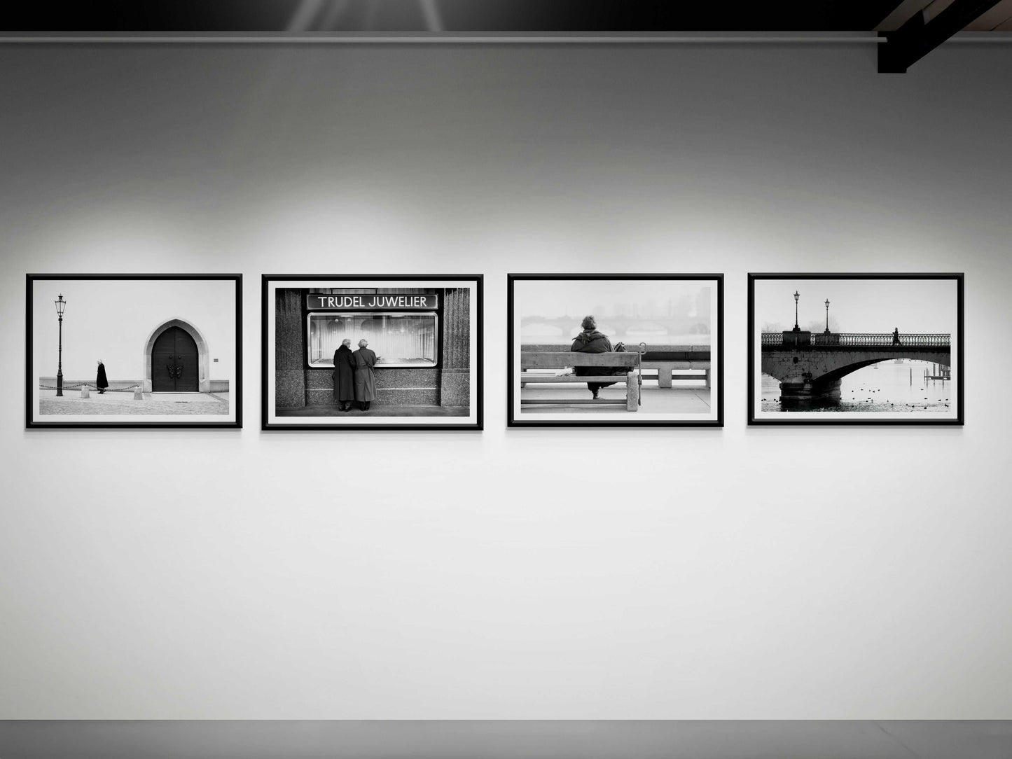 Zurich Series #6 – The Woman Overlooking the River Limmat fine art print by Michael Hart displayed in a gallery with other black and white European street scenes. A striking addition to collectible photography exhibits.